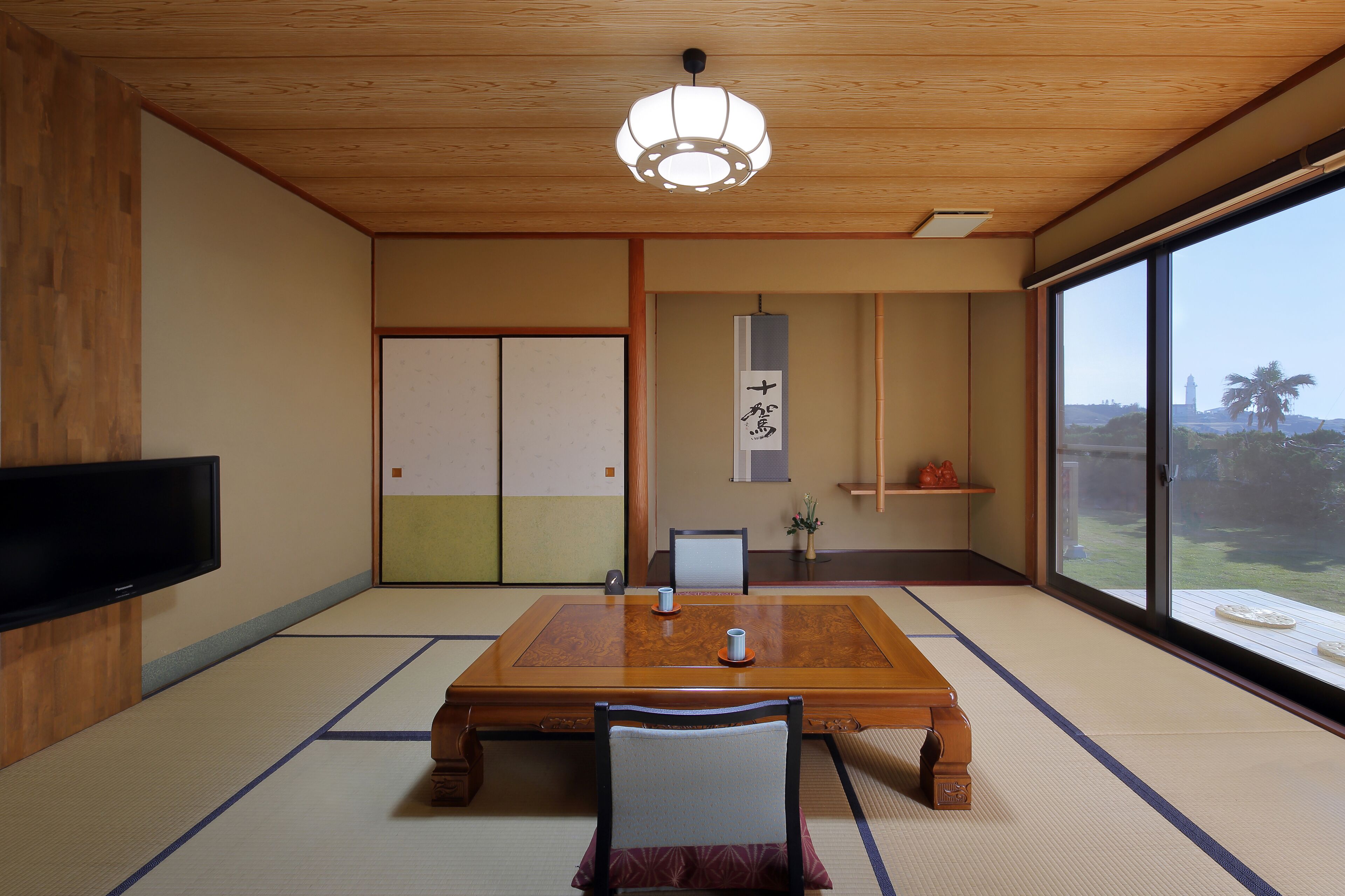 A traditional Japanese-style room with tatami flooring, a wooden table, a television mounted on the wall, and a sliding door.