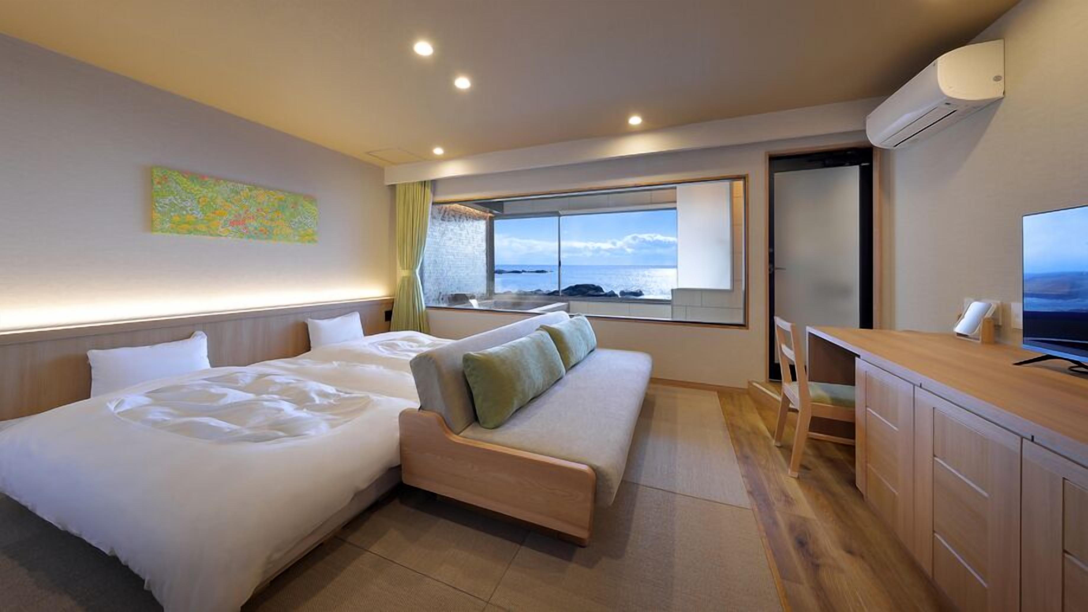 A hotel room with two beds, a sofa, a TV, and a window with a view of the sea.