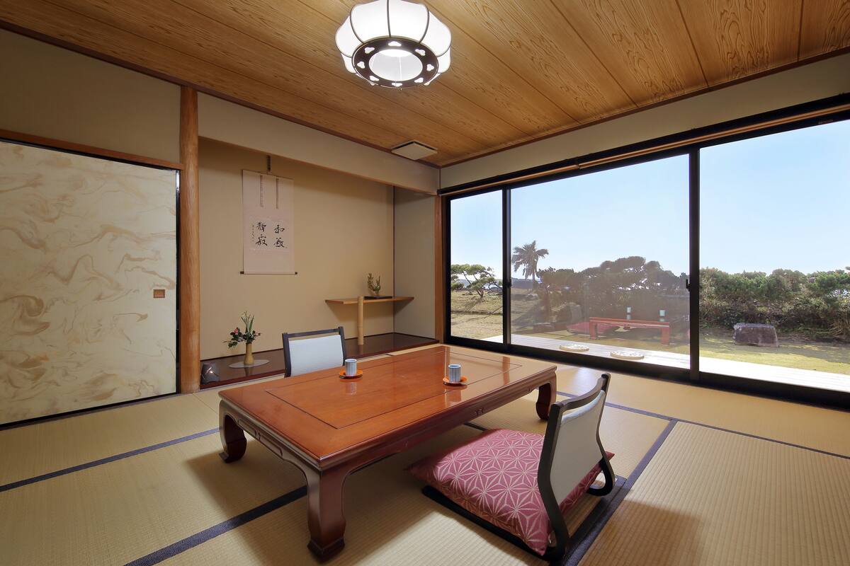 Une pièce de style japonais traditionnel, avec un sol recouvert de tatamis, une table en bois, des tapis de tatami, un paravent shoji et une vue sur l’extérieur.
