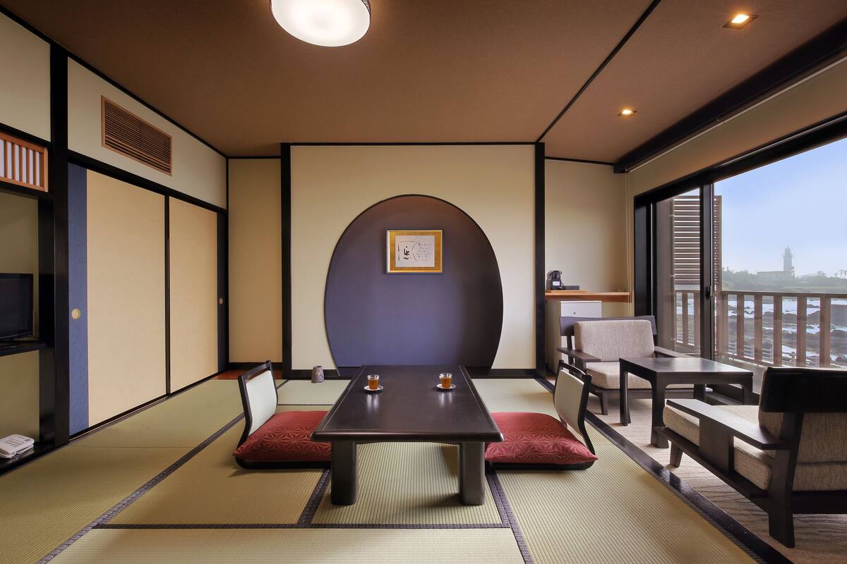 Une pièce de style japonais traditionnel, avec un sol recouvert de tatamis, une petite table sur laquelle sont posées deux tasses, et une vue sur le paysage urbain à travers de grandes fenêtres.