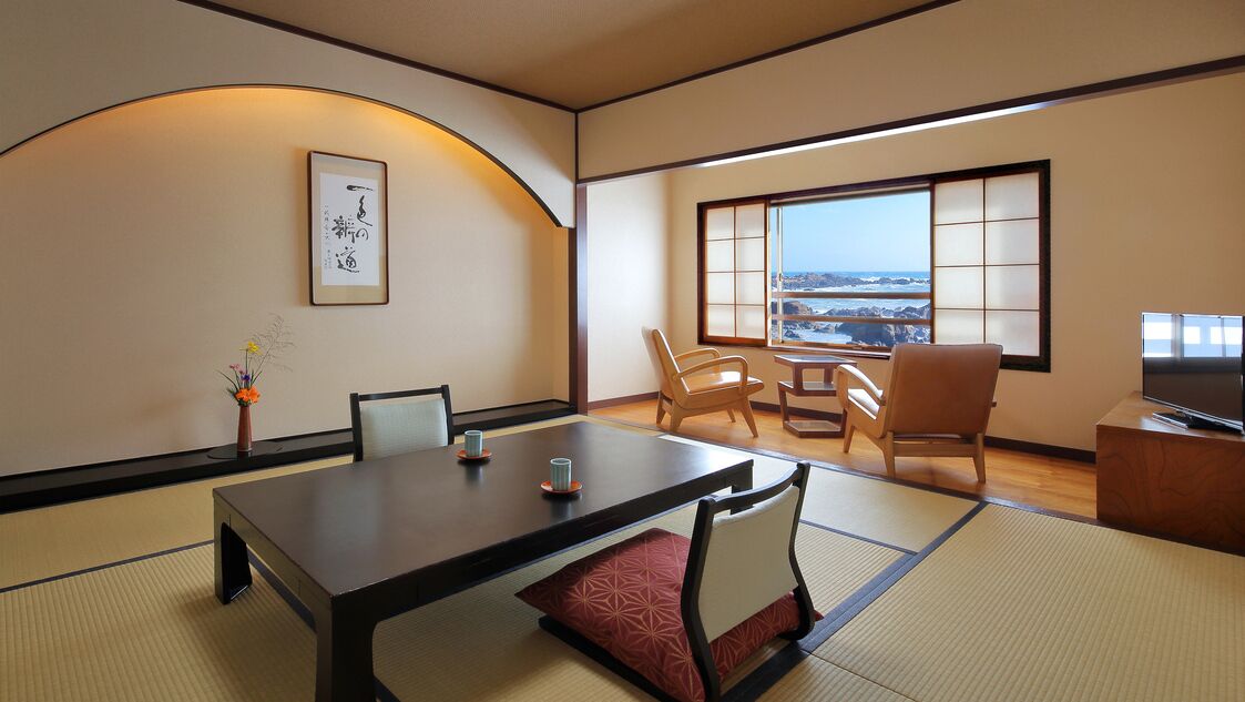 A traditional Japanese room with tatami flooring, a low table with chairs, a TV, and a large window with a view.