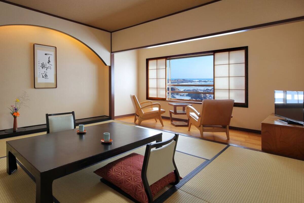 Une pièce de style japonais traditionnel, avec un sol recouvert de tatamis, une table basse entourée de chaises, un téléviseur et une grande fenêtre offrant une belle vue.