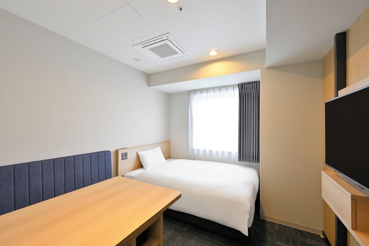 A hotel room with a bed, a wooden desk, a flat-screen TV, and a window with curtains.