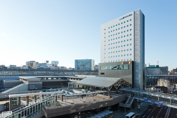 A modern hotel with a prominent tower, a covered parking area, and a train station in the background.