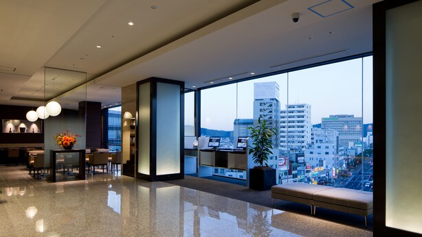 A modern hotel lobby with a large window offering a city view, a reception desk, seating area, and a potted plant. - JR WEST GROUP VIA INN OKAYAMA (Okayama)