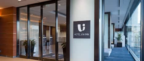 A modern hotel lobby with glass doors, a reception desk, and a staircase leading to upper floors.