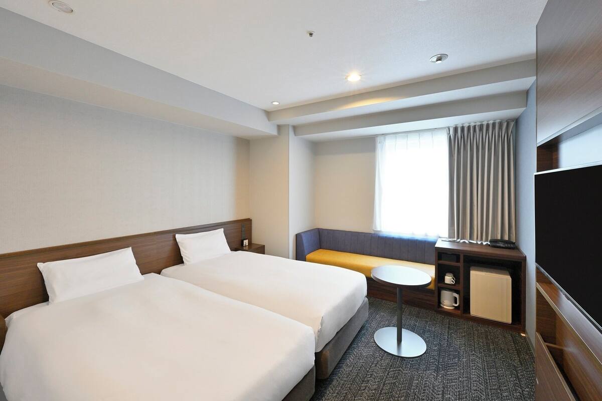 A hotel room with two beds, a small table, a sofa, a TV, and a window with curtains.