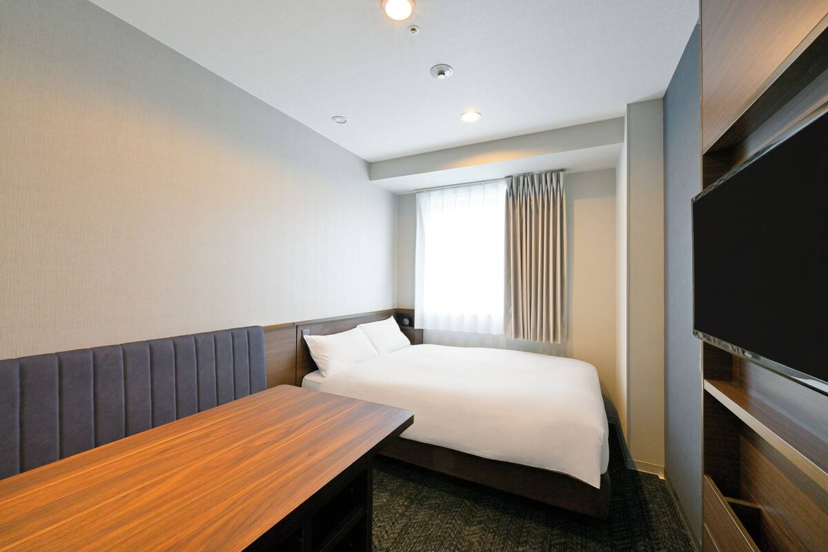 A hotel room with a bed, a wooden bench, a flat-screen TV, and a window with curtains.
