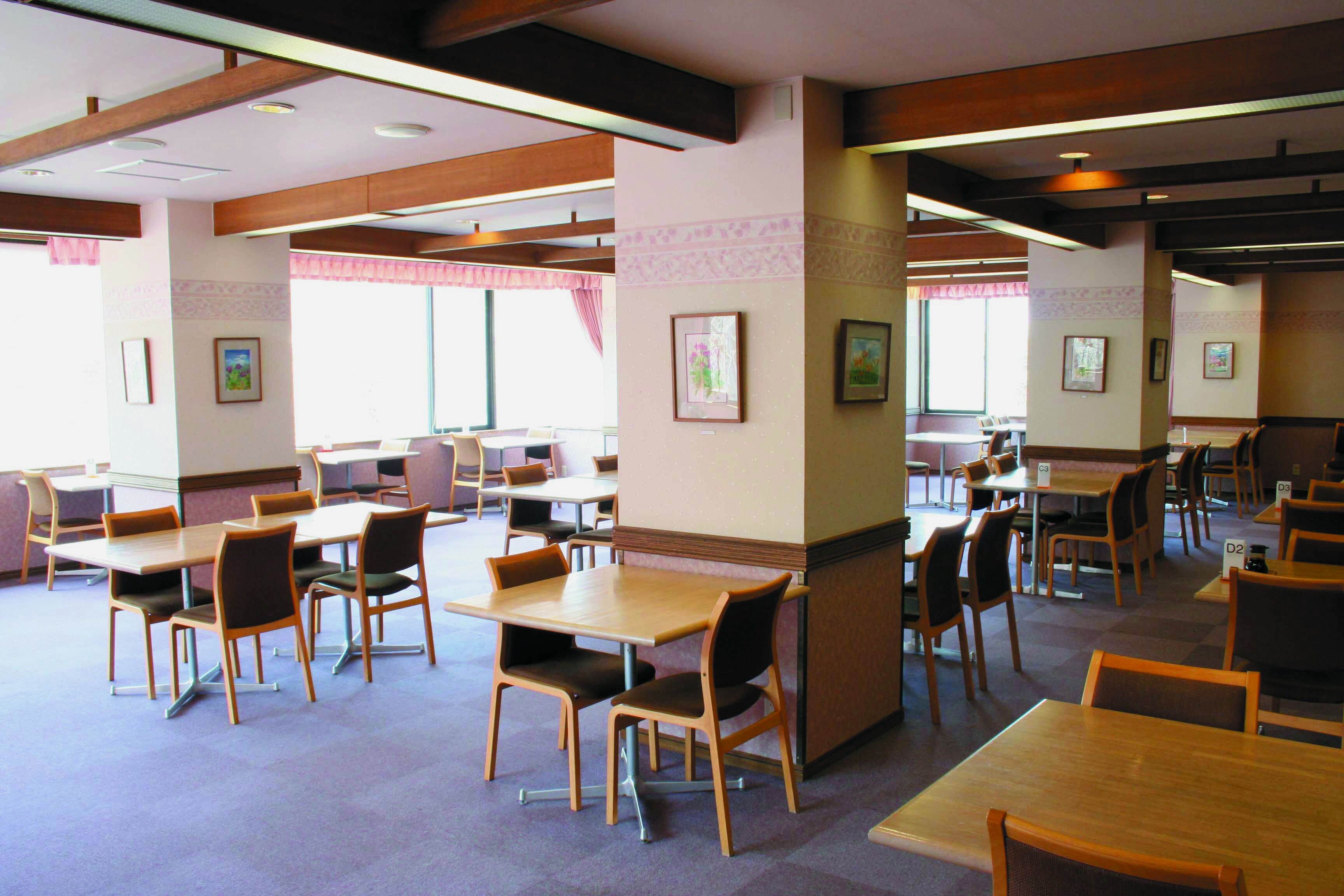 Un'ampia sala da pranzo interna con numerosi tavoli e sedie, ampie finestre con tende e quadri appesi alle pareti.