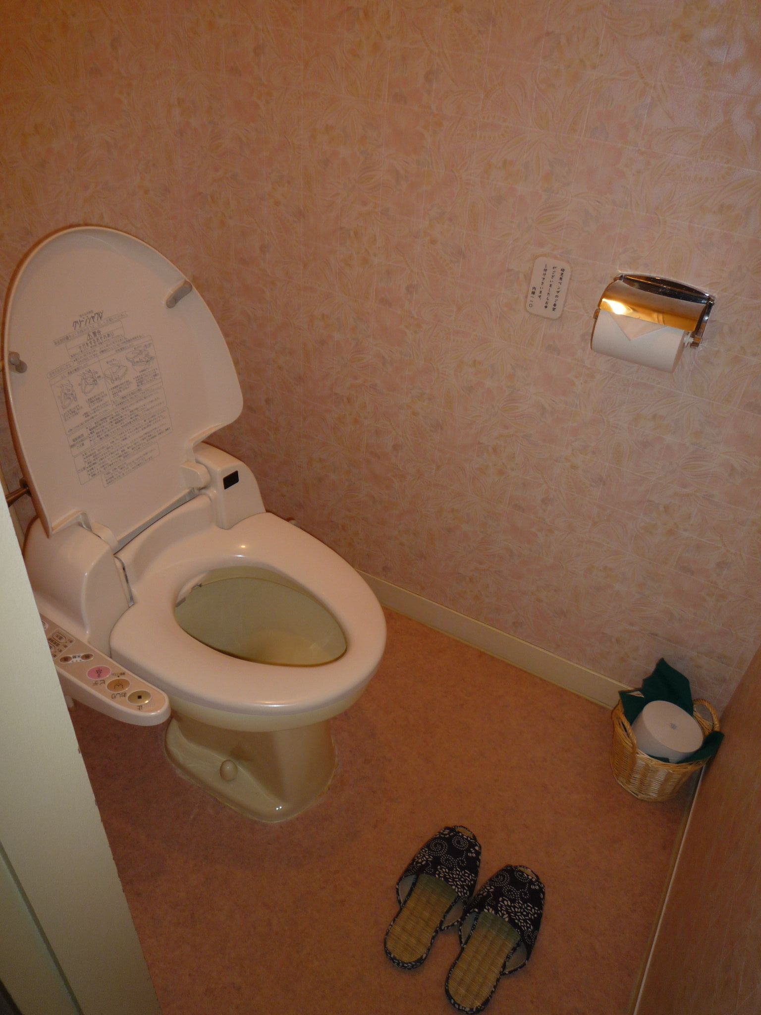 Un baño con un inodoro, un dispensador de toallas de papel y un par de pantuflas en el suelo.