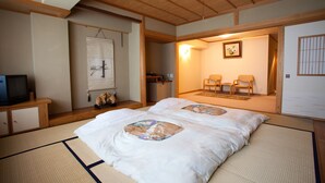 Una habitación tradicional japonesa con suelo de tatami, una cama con colcha de estampado floral, un televisor sobre un mueble de madera y una puerta corredera que da a una zona de estar con sillas y un cuadro enmarcado.