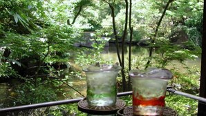 Tres vasos de bebidas heladas sobre una mesa, con una baranda metálica que da a una masa de agua y follaje.