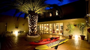 A well-lit outdoor area with a palm tree, a red kayak, and a yellow paddleboard. - Hotel Familio Tateyama (Tateyama)
