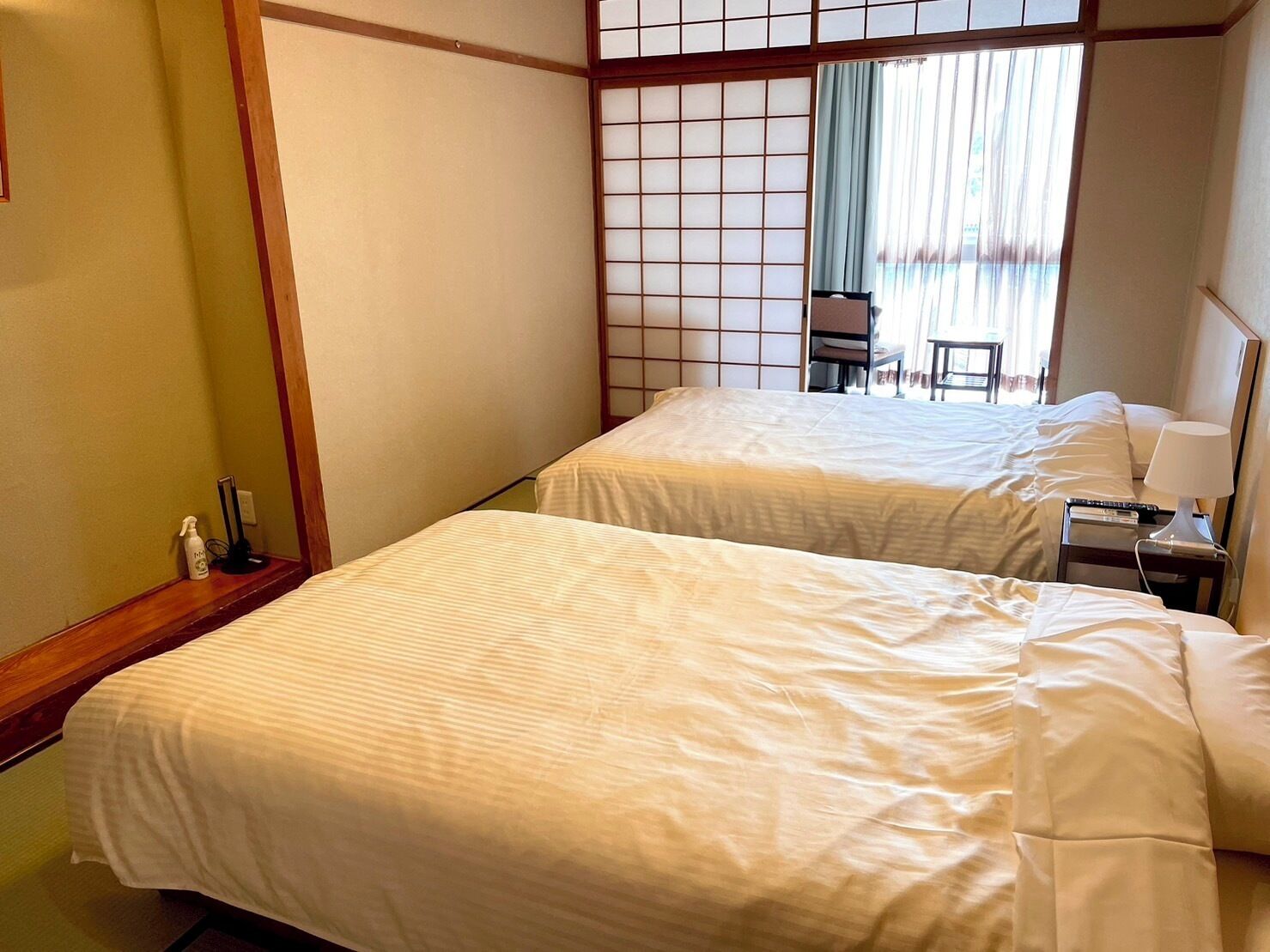Hotelkamer met twee bedden, een houten vloer, een shoji-scherm, een bureau met lamp en uitzicht buiten door een raam met gordijnen.