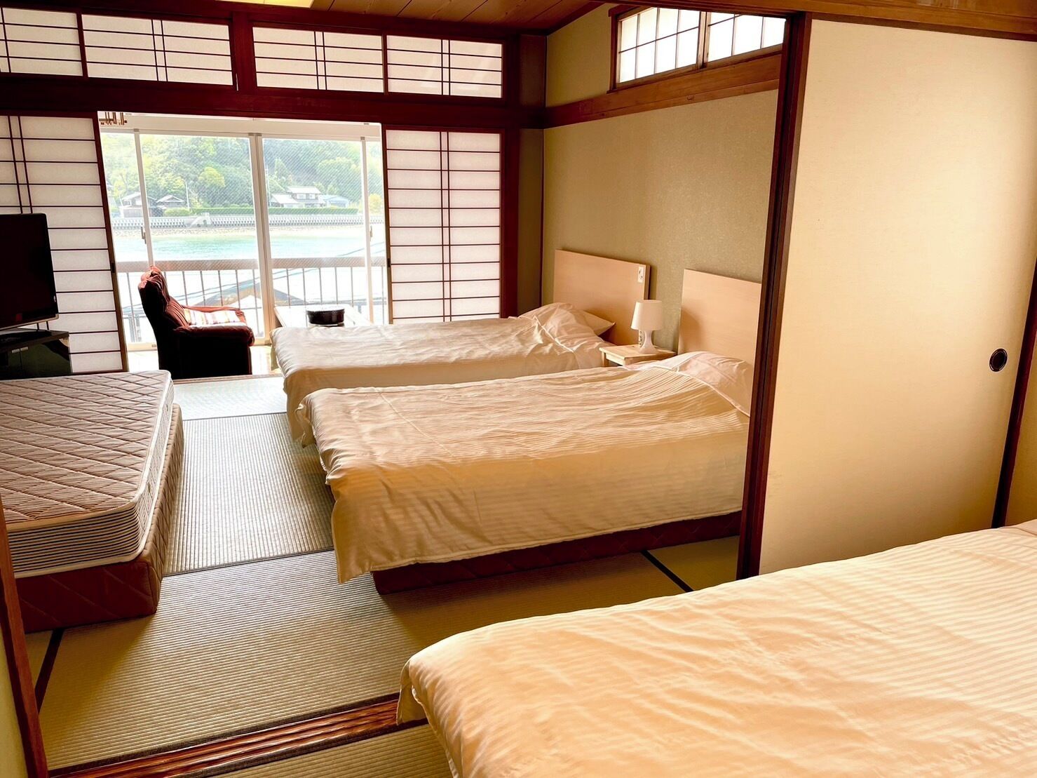 Een traditionele Japanse hotelkamer met tatami vloerbedekking, twee bedden, een televisietoestel en een schuifdeur naar een balkon met uitzicht op een wateroppervlak.