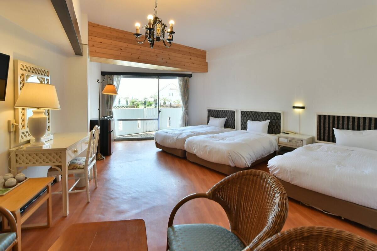 A hotel room with two beds, a desk with a lamp, a chair, a TV, and a balcony with a view.