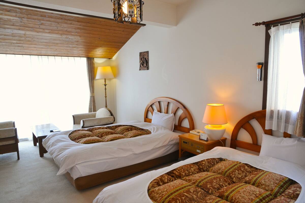 A hotel room with two beds, a wooden headboard, bedside lamps, a small table, and a chair.