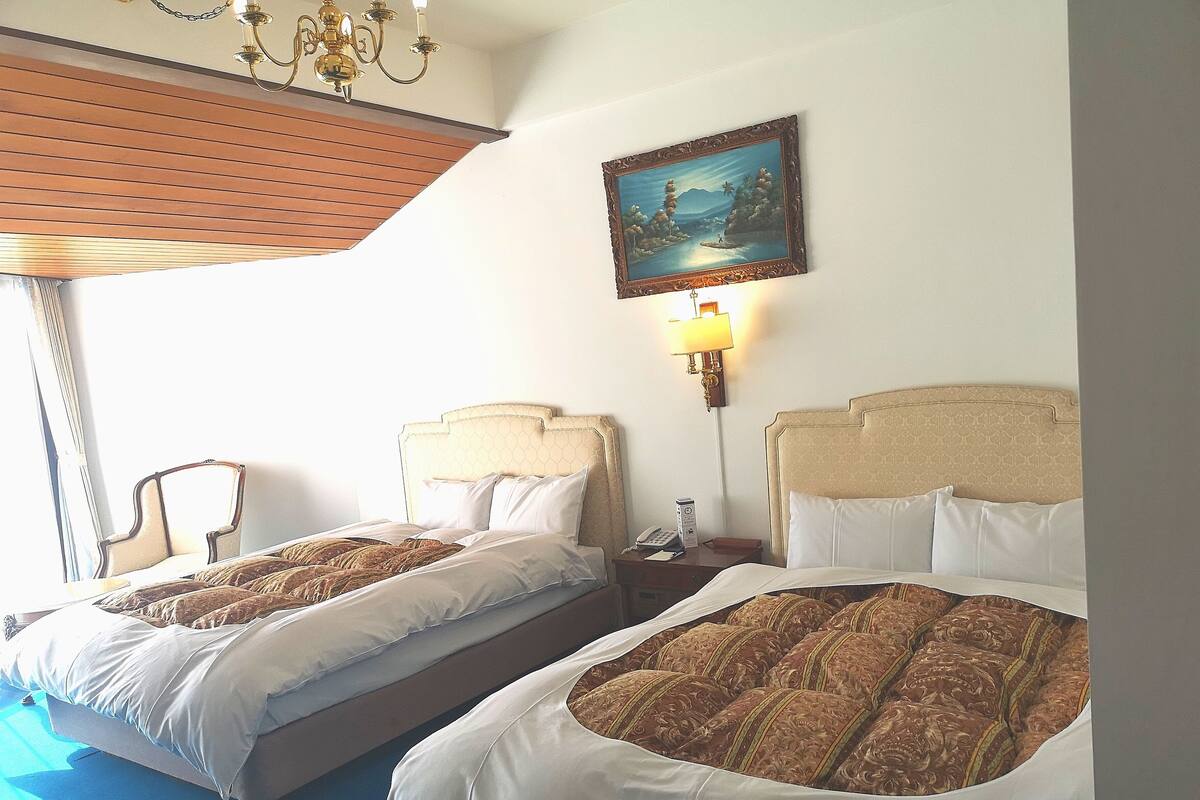 A hotel room with two beds, a chandelier, a painting, and a bedside table with a lamp.