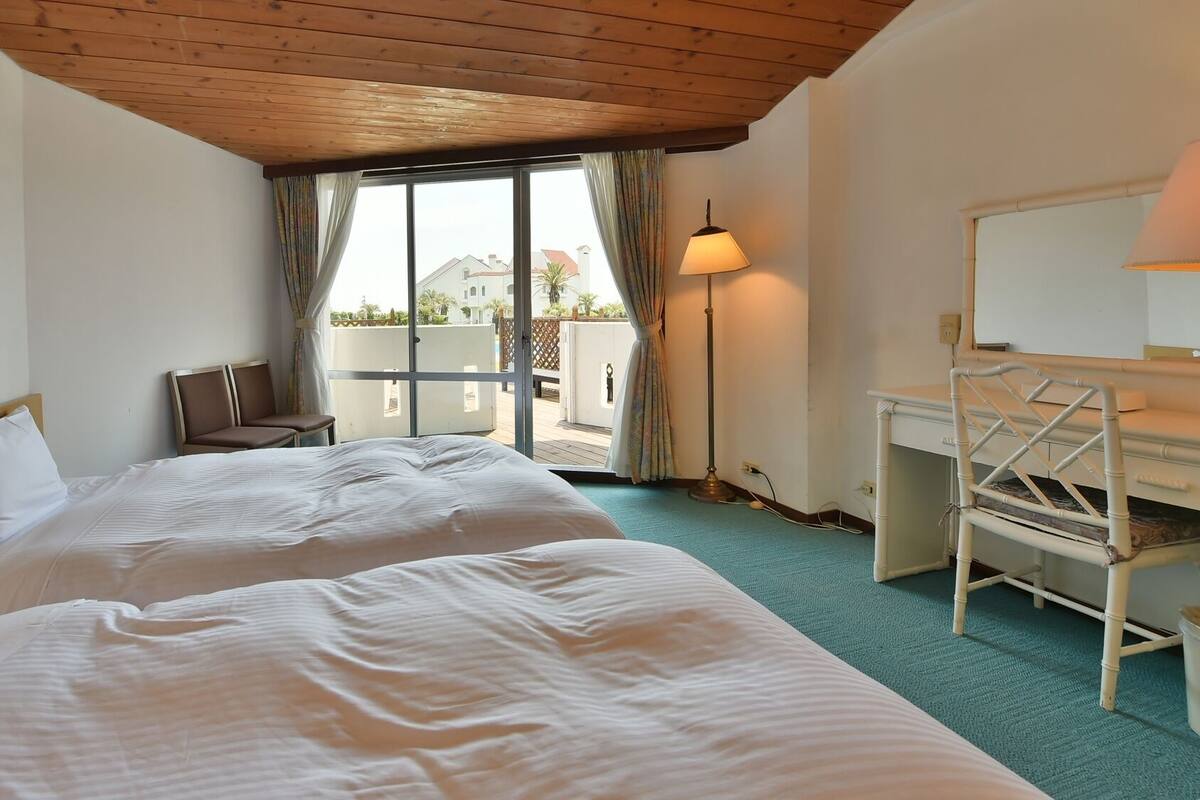 A hotel room with two beds, a desk, a chair, and a balcony with a view.
