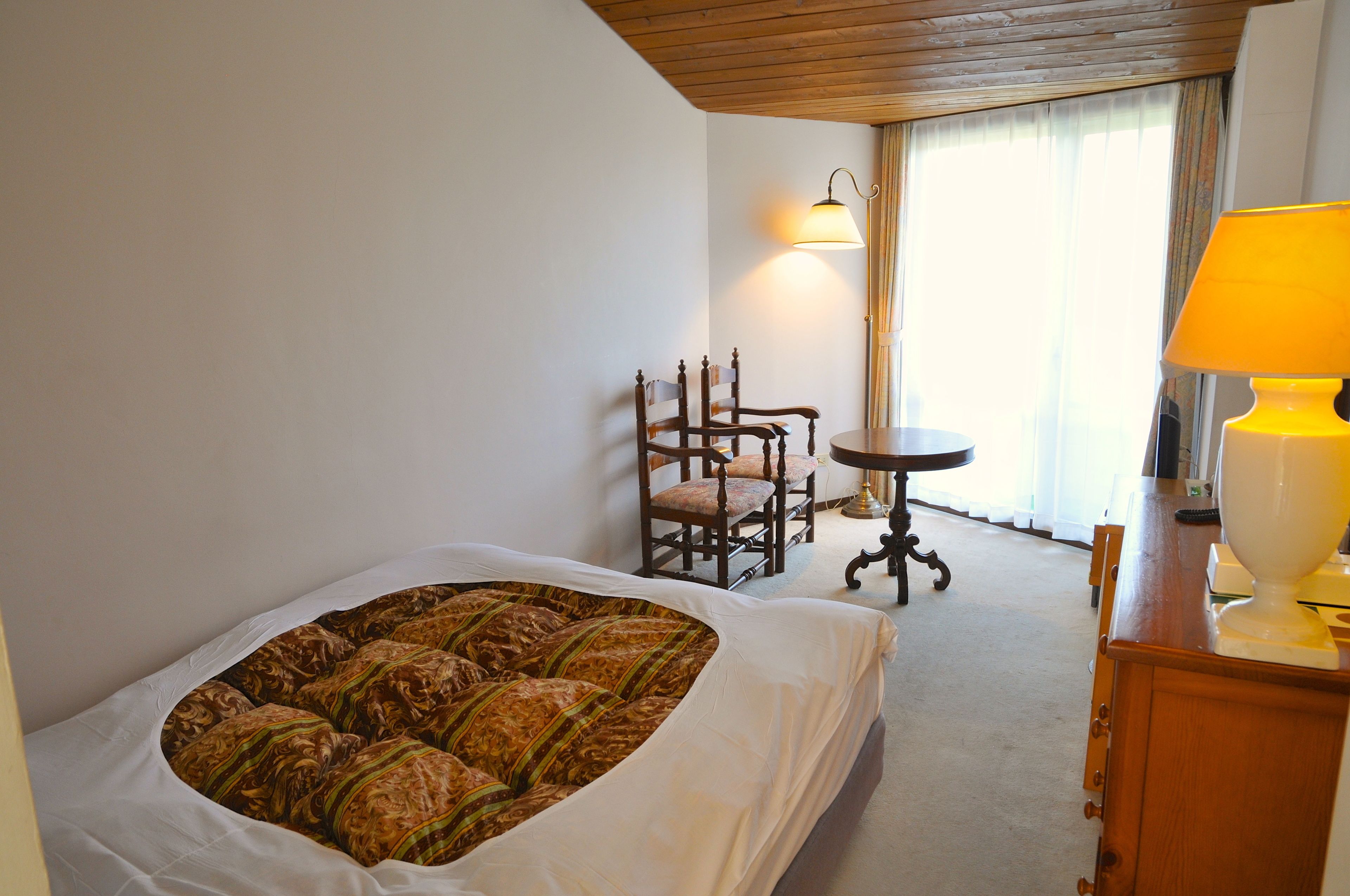 A hotel room with a bed, a wooden chair, a small table, and a lamp.