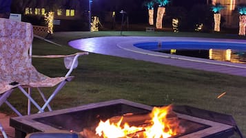 A night scene with a fire pit, a chair, and a pool with lights.
