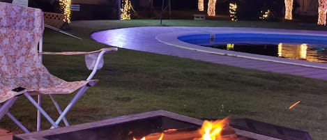 Escena nocturna con una fogata, una silla y una piscina iluminada.