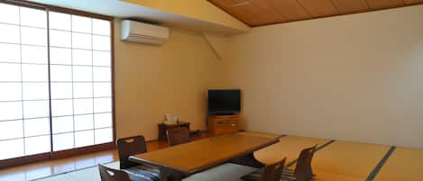 Um quarto japonês tradicional com tatames, uma mesa baixa, almofadas, uma televisão e portas deslizantes.