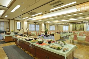 Uma ampla sala de banquetes com várias mesas preparadas para refeições, um buffet e uma grande janela que proporciona iluminação natural.