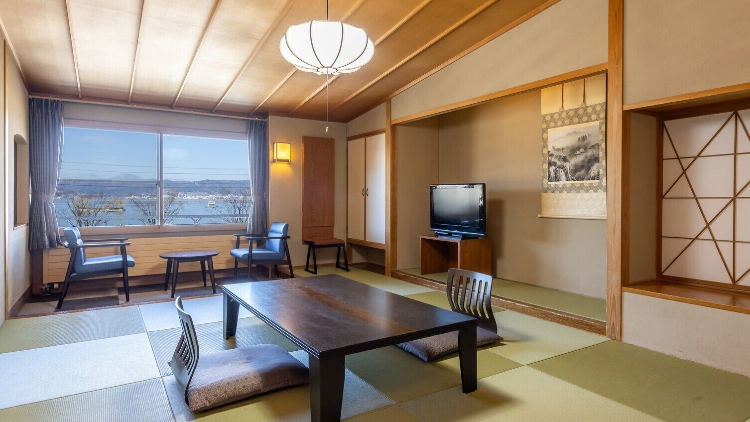 A traditional Japanese room with tatami flooring, a low table, chairs, a television, and a large window offering a view.