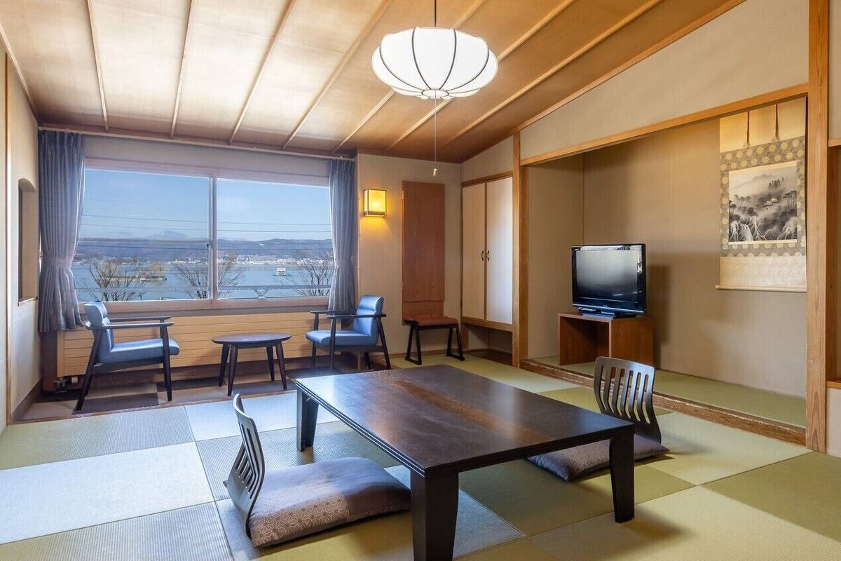 Una habitación japonesa tradicional con suelo de tatami, una mesa baja, sillas, un televisor y un ventanal con vistas amplias.