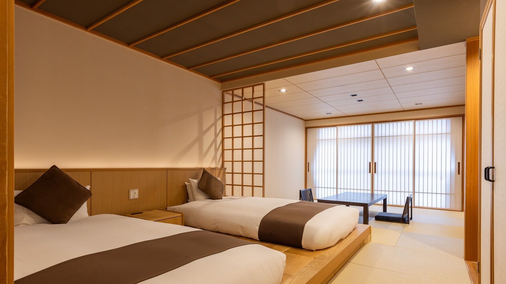 A hotel room with two beds, a sliding door, a table, and a view of the outside.