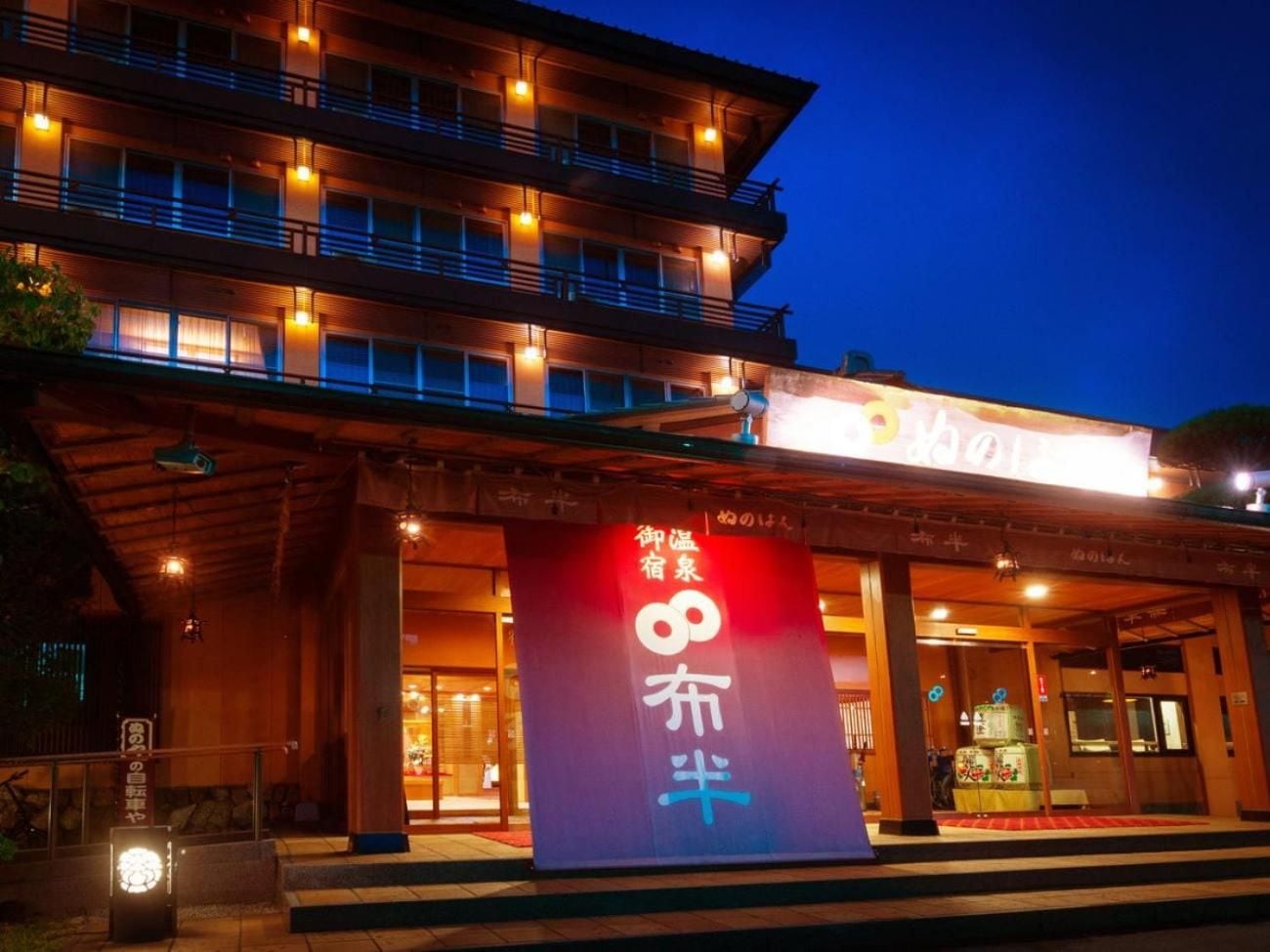 A hotel entrance with a large sign displaying Japanese characters, a covered walkway, and a multi-story building with illuminated windows.