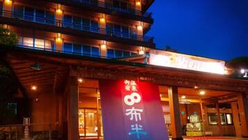 A hotel entrance with a large sign displaying Japanese characters, a covered walkway, and a multi-story building with illuminated windows.