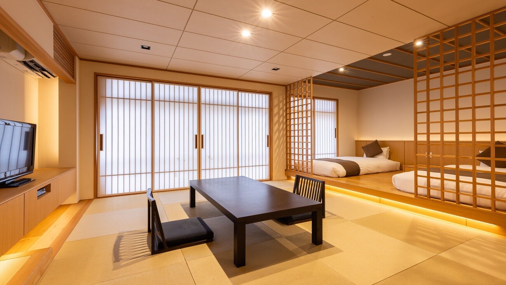 A traditional Japanese room with tatami flooring, a low table with a bench, a television, sliding doors, and a bed with a low headboard.