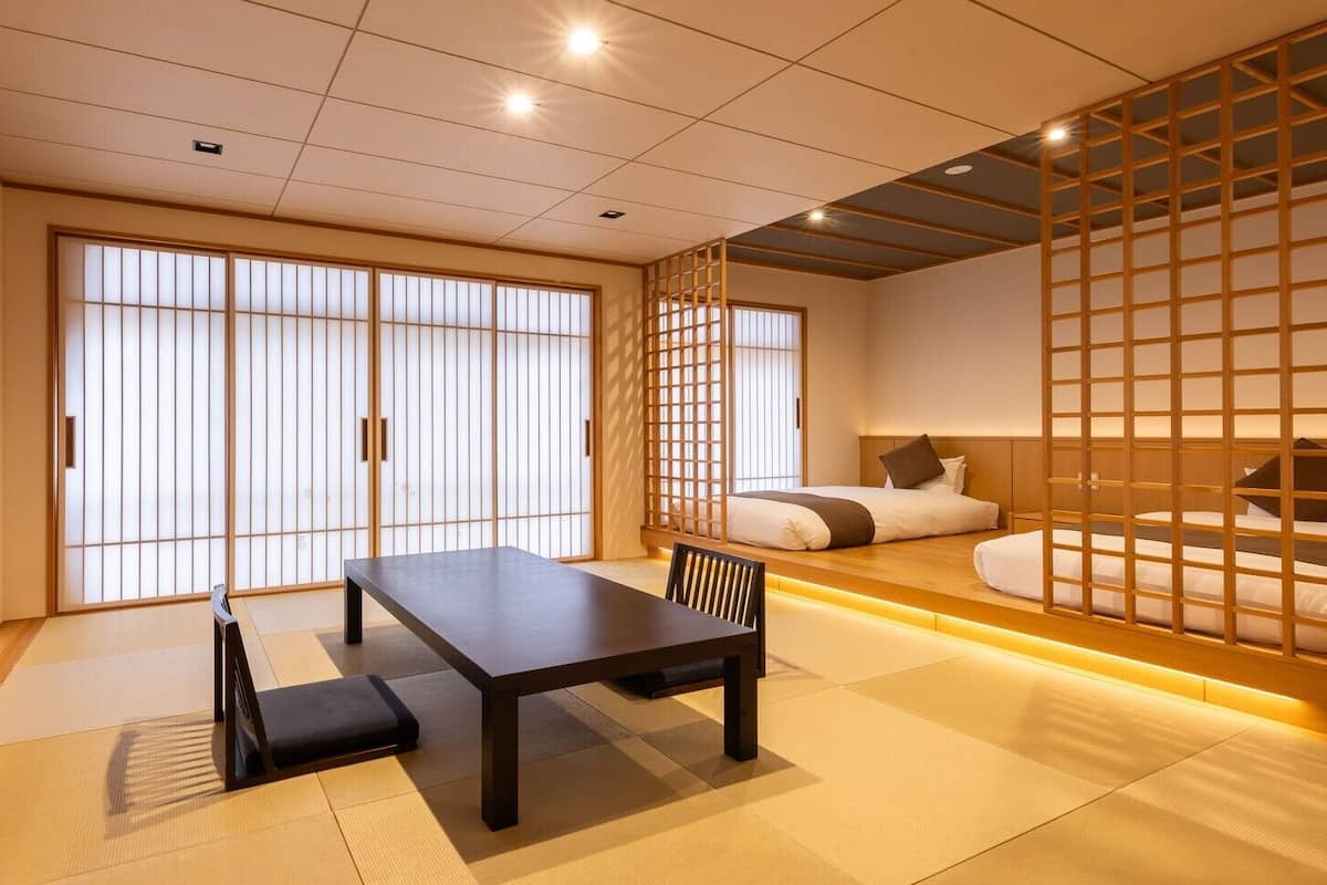 Una habitación japonesa tradicional con suelo de tatami, una mesa baja con un banco, un televisor, puertas correderas y una cama con cabecero bajo.