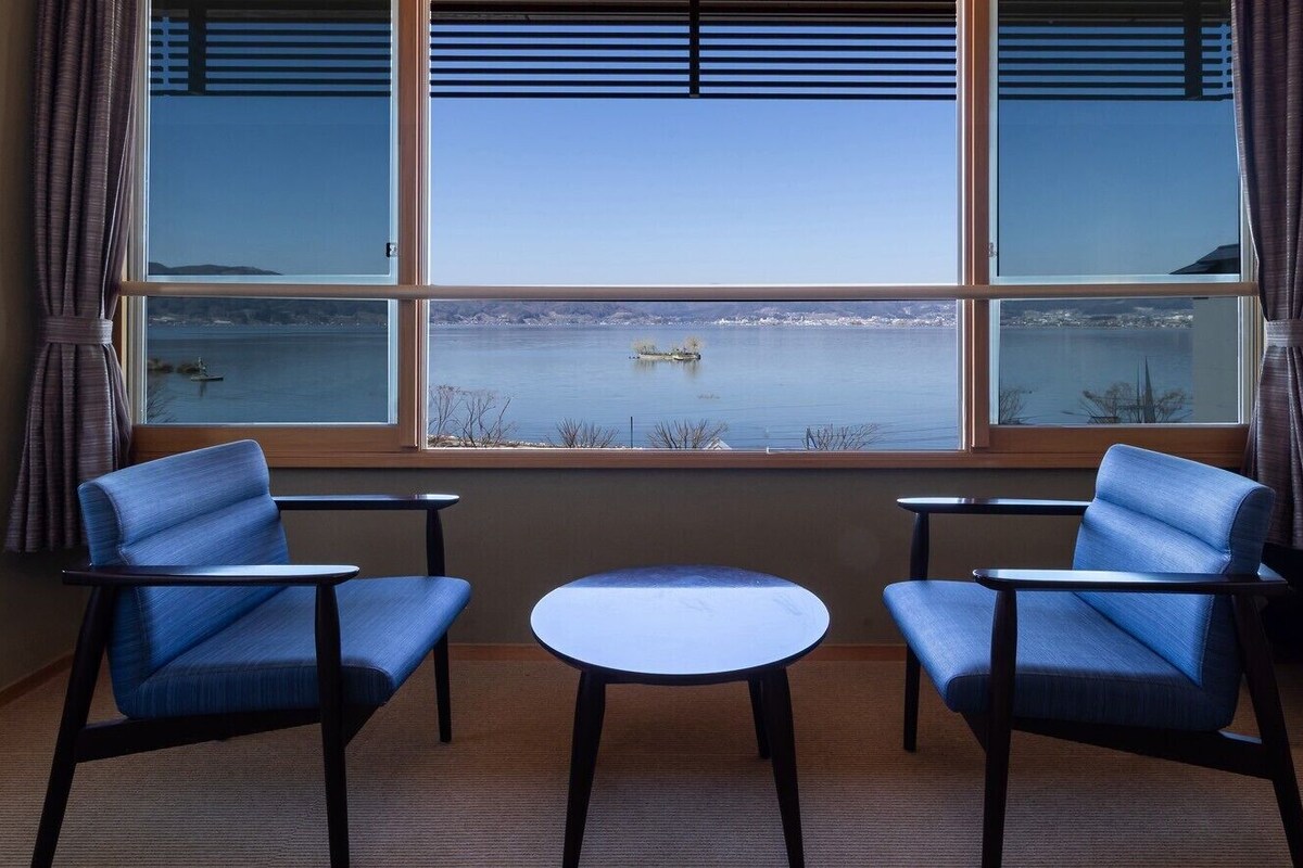 Ett rum med två blå stolar och ett litet bord, ett stort fönster med utsikt över en sjö eller ett hav och ett avlägset landområde.