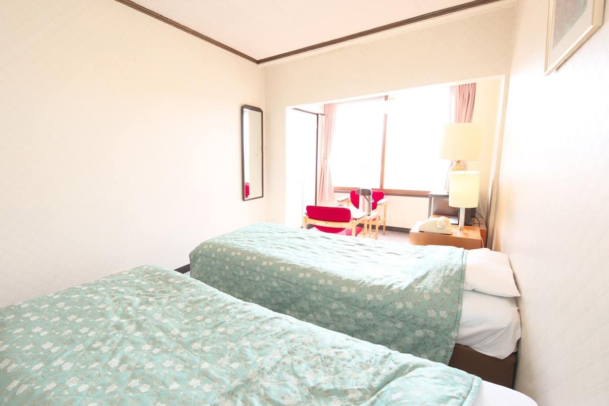 Ein Hotelzimmer mit zwei Betten, einem Schreibtisch, einem Stuhl, einer Lampe und einem fenster mit Vorhängen.