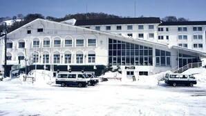 Un hotel con un amplio frente de vidrio, una escalera que conduce a la entrada y un estacionamiento cubierto de nieve.