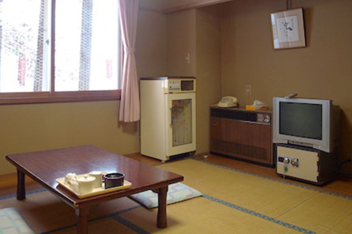 A room with a tatami mat floor, a wooden table with cups and a small container, a refrigerator, a microwave, a telephone, and a television.