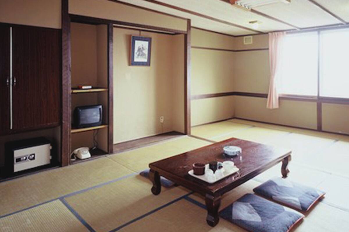 Tatami-mattoilla päällystetty huone, jossa on tatami-matto lattiana, puinen pöytä, jonka päällä on teetarjoilu, hyllyllä oleva televisio ja liukuovi.
