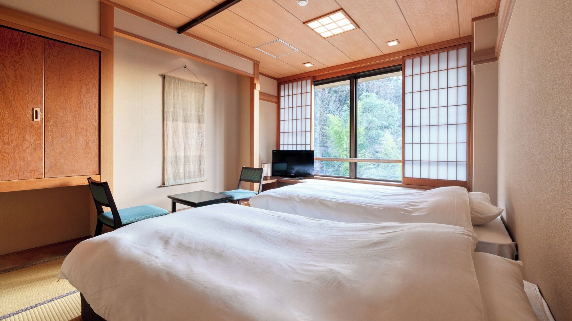 A hotel room with a large bed, a TV, a desk, and a view of trees through the window.