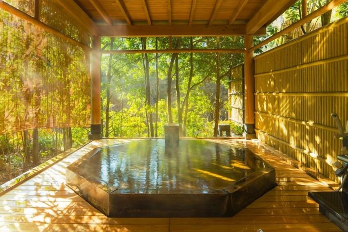 Un área de baños termales con una terraza de madera, rodeada de paredes de bambú y árboles.