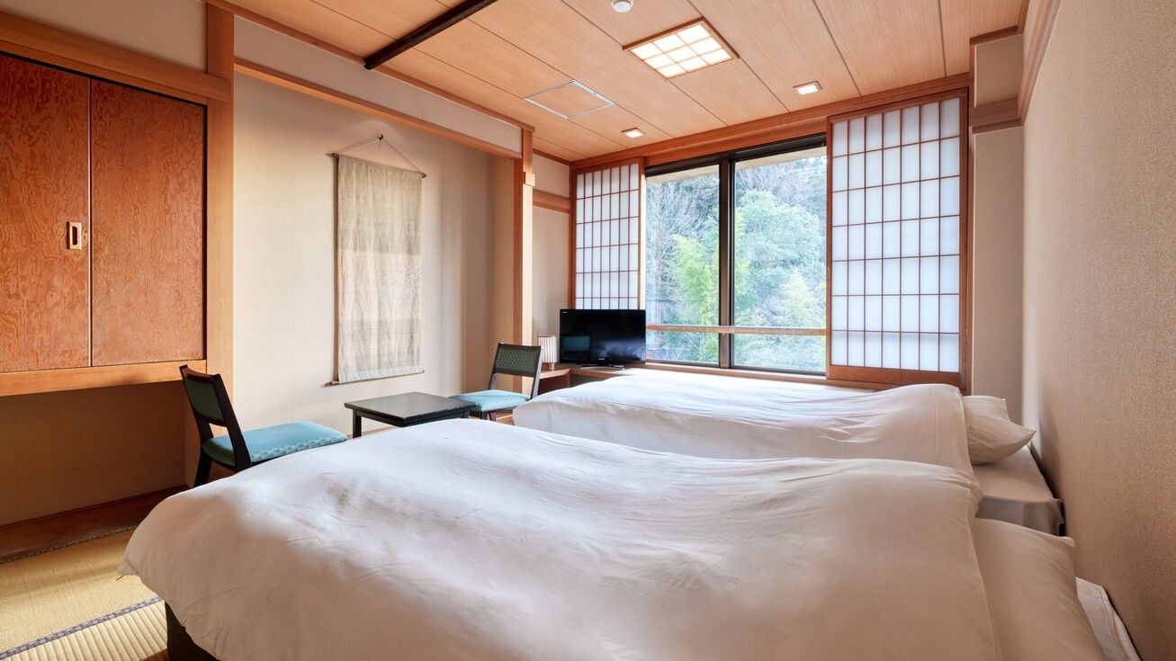 A hotel room with a bed, a television, a desk, and a view of trees through large windows.