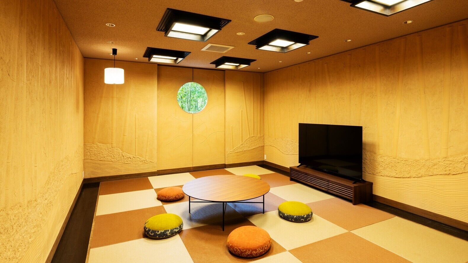 Een kamer met een ronde tafel, vier kleurrijke kussens, een groot scherm en een rond raam met uitzicht op bomen.