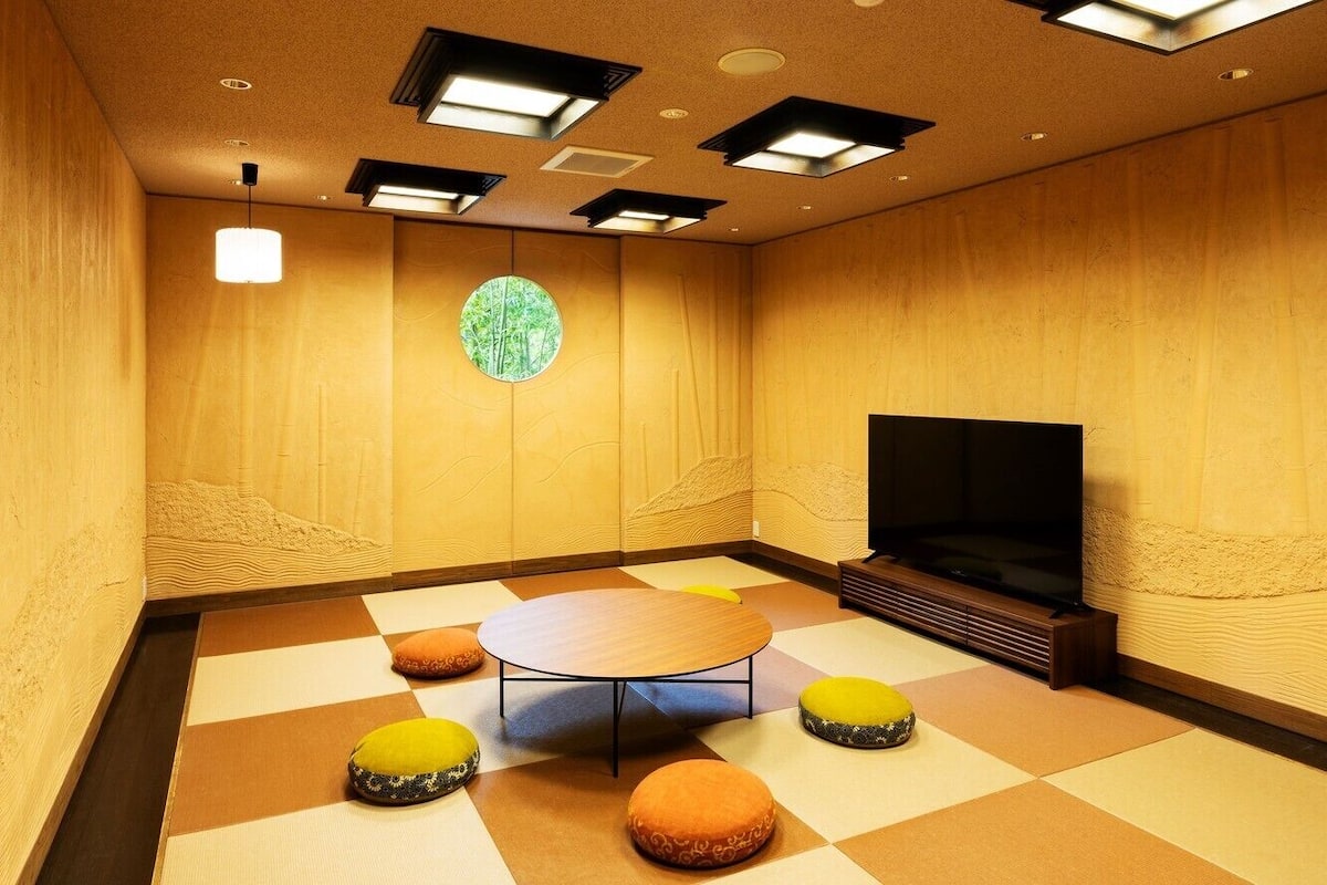 Une pièce avec une table ronde, quatre coussins colorés, un grand écran et une fenêtre circulaire donnant sur la forêt.