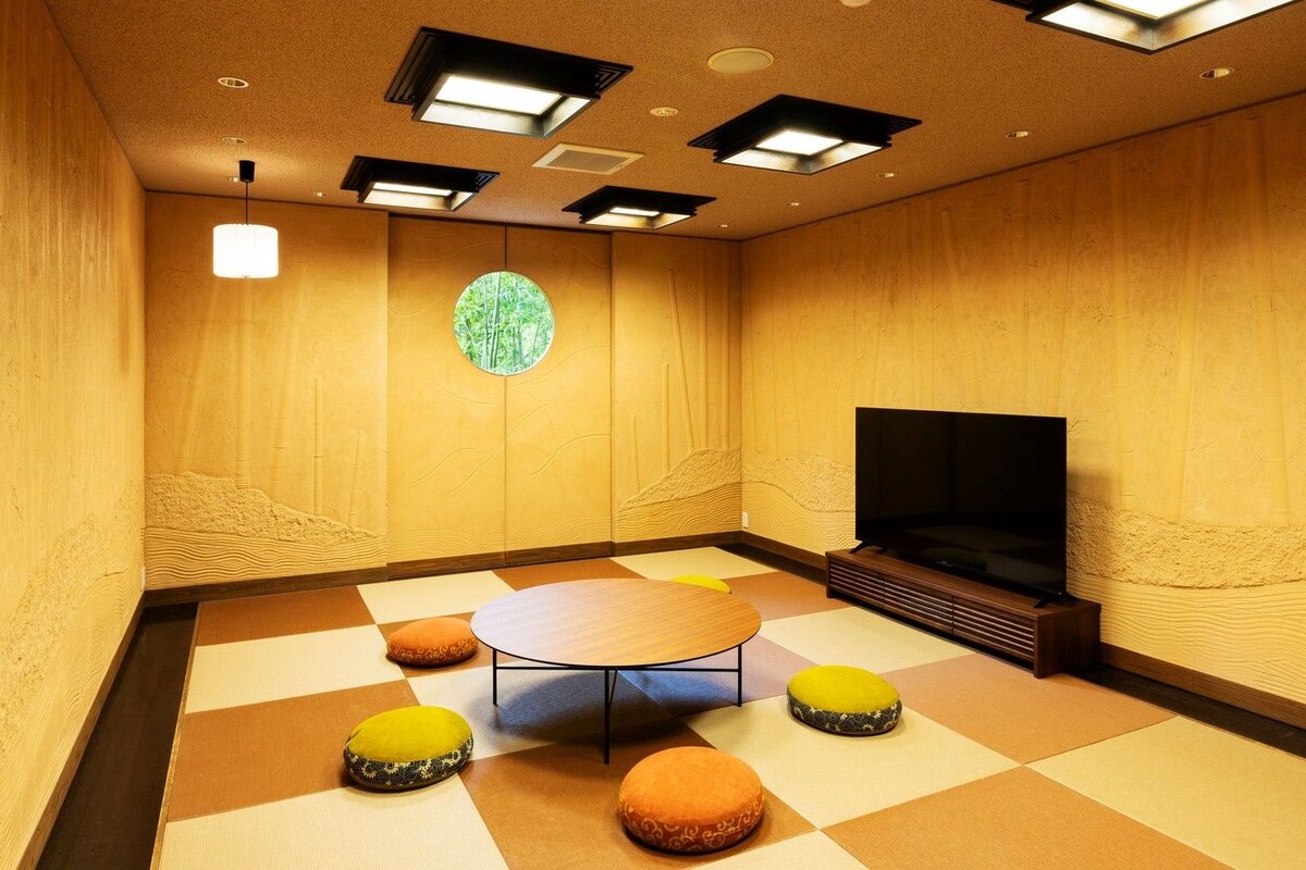 Una habitación con una mesa redonda, cuatro cojines de colores, una pantalla grande y un ventanal circular con vistas a árboles.
