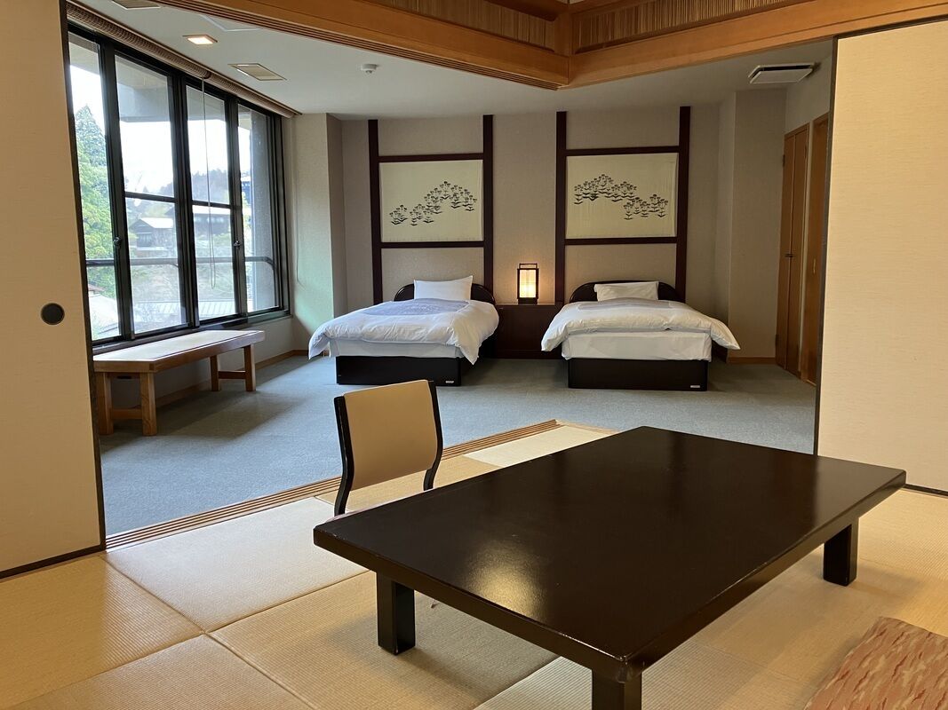 Een hotelkamer met twee bedden, een houten tafel, tatami vloeren en grote ramen.
