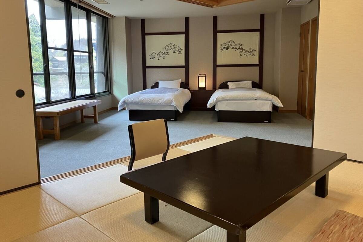 Une chambre d’hôtel avec deux lits, une table en bois, un sol recouvert de tatamis et de grandes fenêtres.
