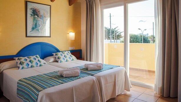 A hotel room with two beds, a large window with curtains, a painting on the wall, and a view of the outside. - Sagitario Villas (Ciutadella de Menorca)