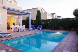 A swimming pool with blue lounge chairs, a covered patio area, and a building with a beige facade. - Sagitario Villas (Ciutadella de Menorca)